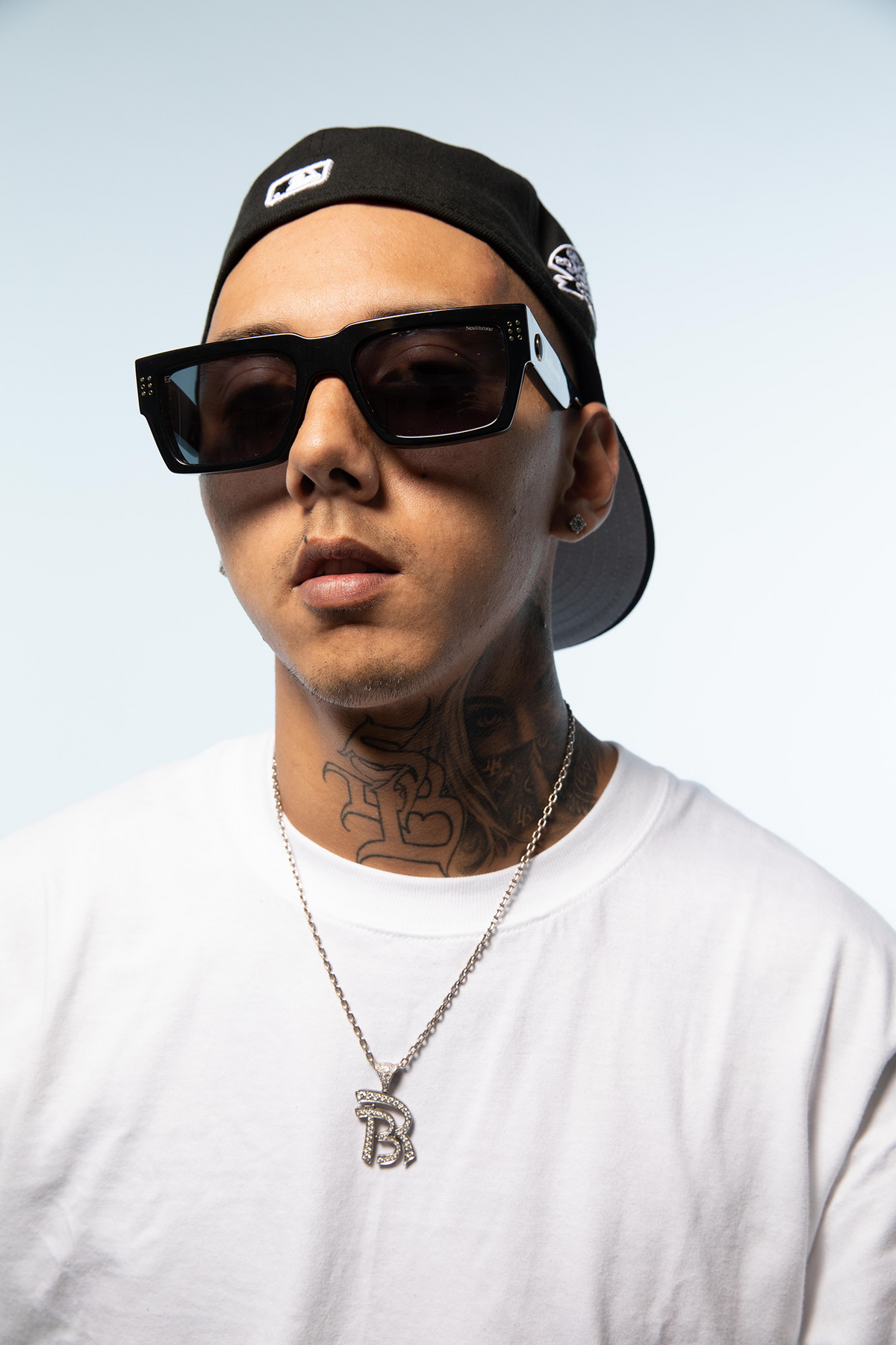 Person wearing sunglasses, a cap, and a white shirt with a necklace against a light background