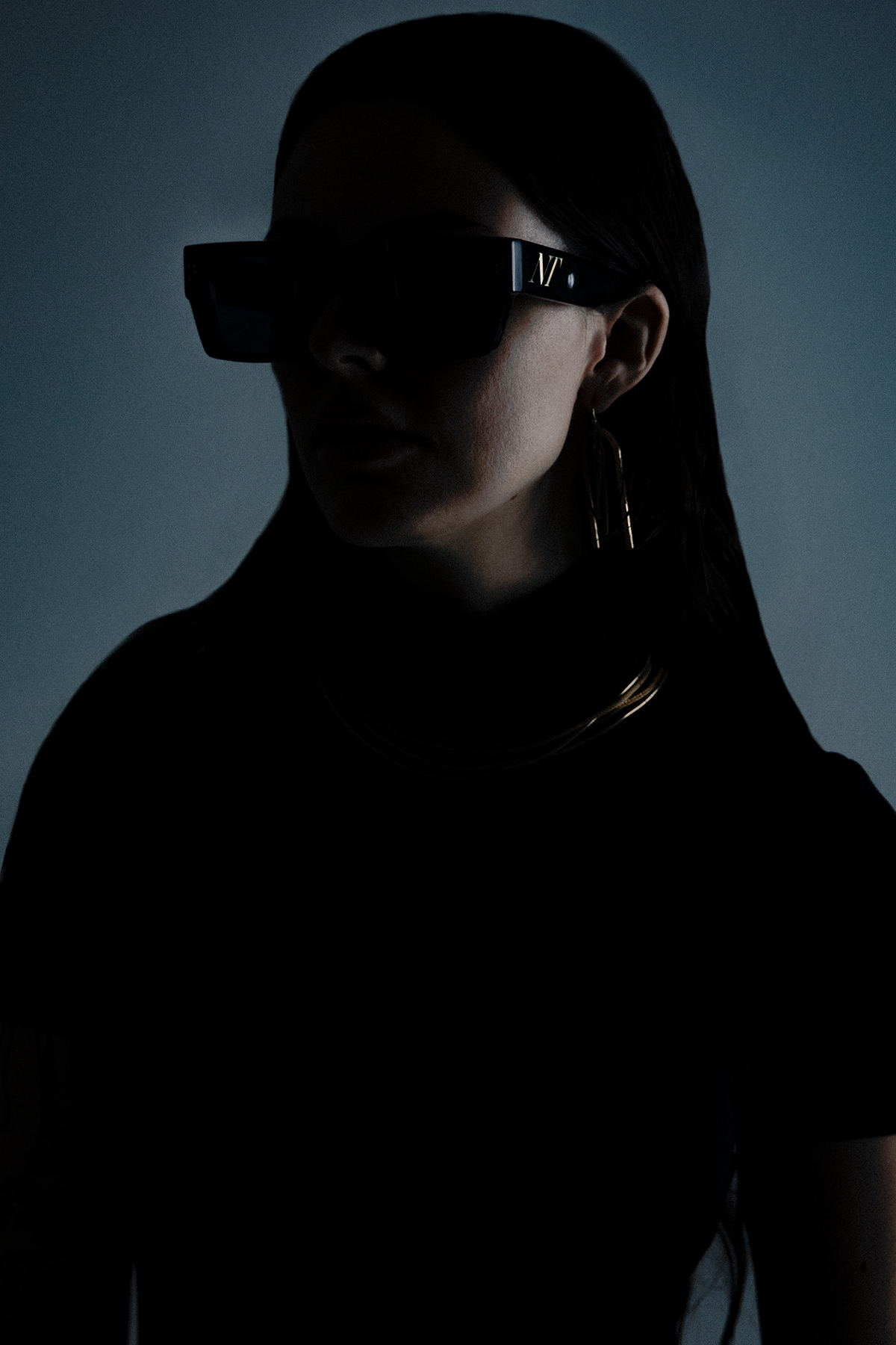 Side portrait of a model wearing black sunglasses and a black top in dim lighting.