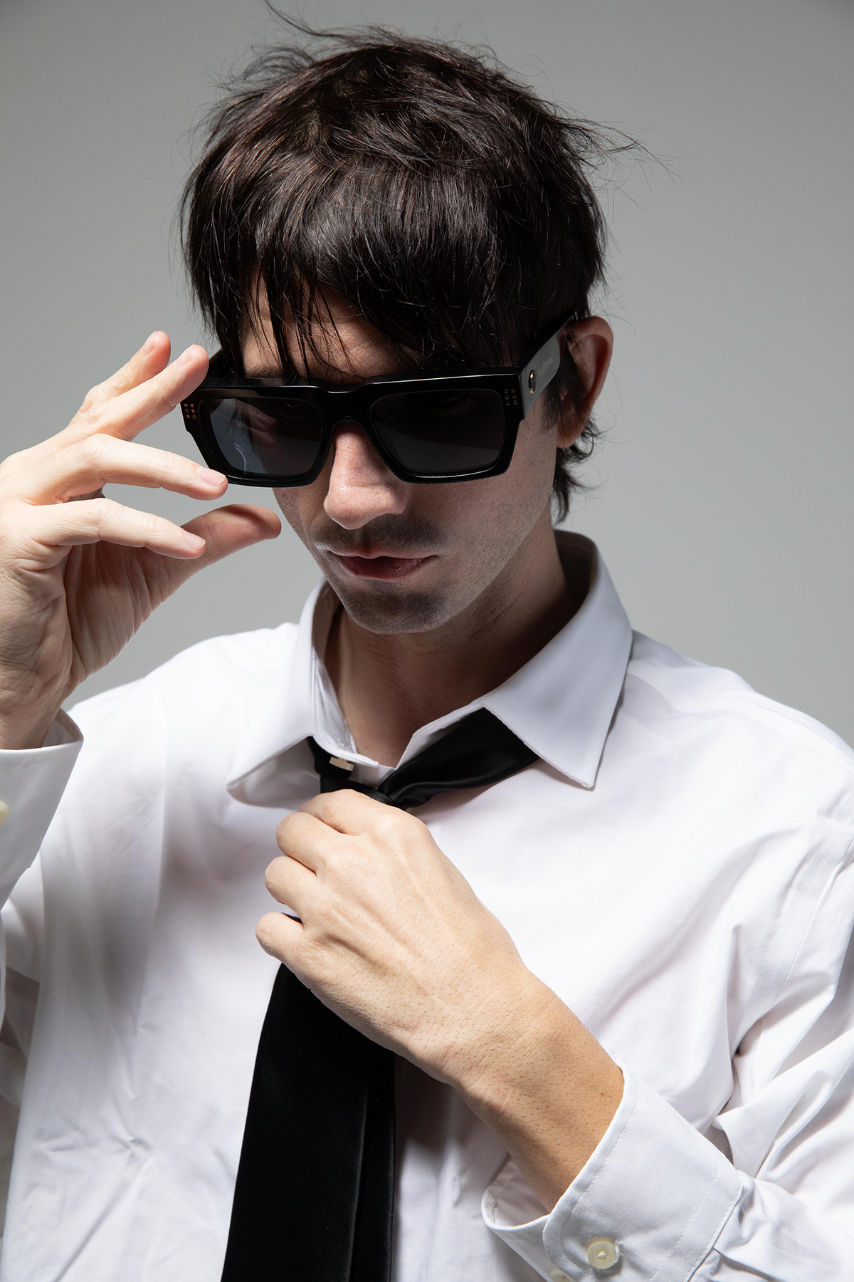 Man wearing sunglasses and adjusting a tie against a gray background