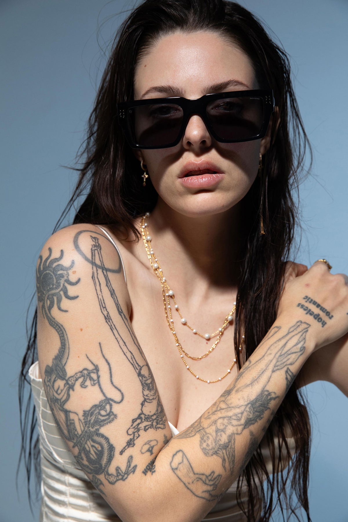 Woman with tattoos wearing sunglasses and layered necklaces against a blue background